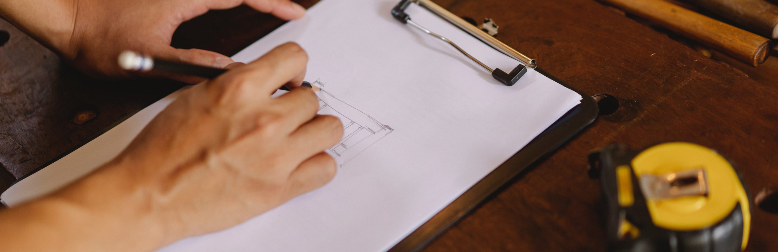 Buildings & Grounds. A close-up of a person’s hand drawing a diagram on a clipboard. A close-up of a person’s hand drawing a diagram on a clipboard. There is a tape measure on the right-hand side of the table.